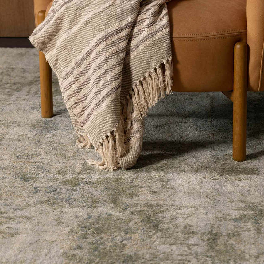 Close-up of the Zazie Thick Luxe Rug in beige, gray, olive, and teal under a chair with a striped throw. Styleshot