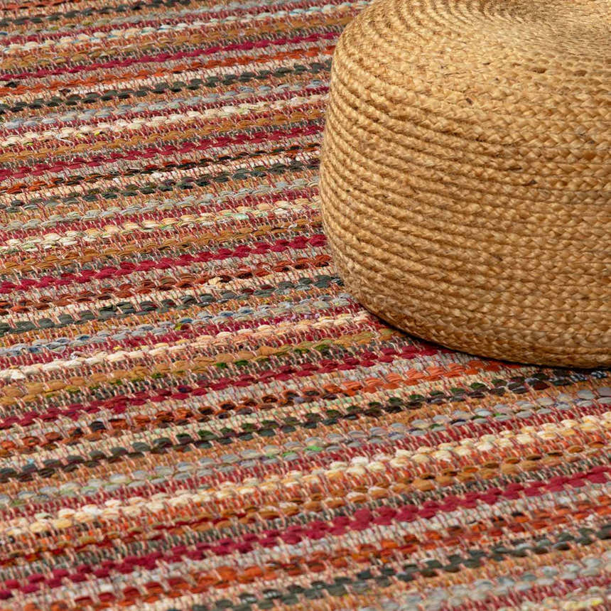 Close-up of the hand woven Brenda Crimson Area Rug with red, orange, and neutral stripes and a jute pouf. Styleshot