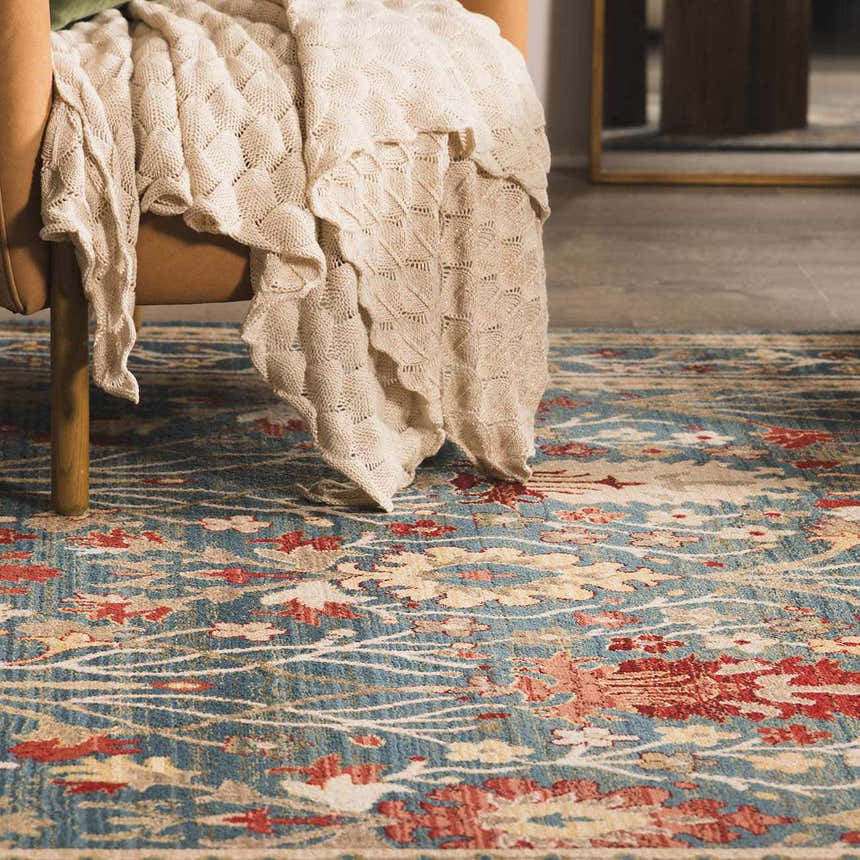 Close-up of the Neola Navy Blue Damask Area Rug featuring a floral pattern in brick red, tan, and white. Styled with a chair and blanket. Roomscene-Rectangle