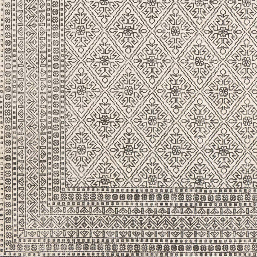 Close-up of the hand-woven Brushton Area Rug. Beige cotton rug with a black geometric diamond and floral pattern. Swatch