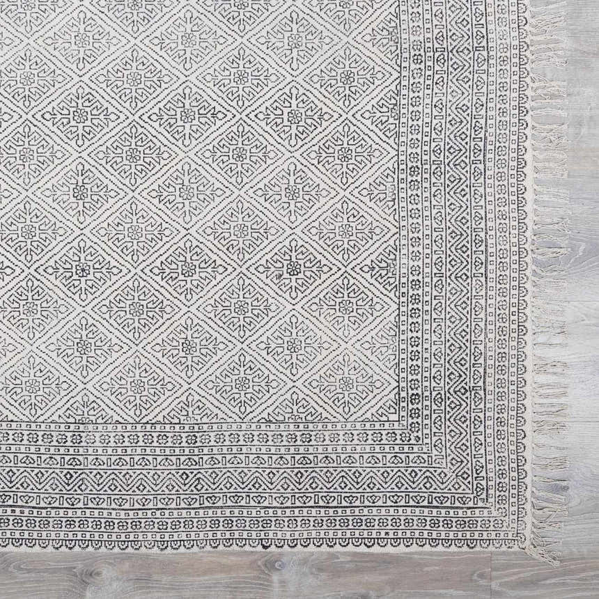 Close-up of the Brushton Area Rug. Beige cotton rug with black geometric diamond and border patterns, and fringe. Swatch