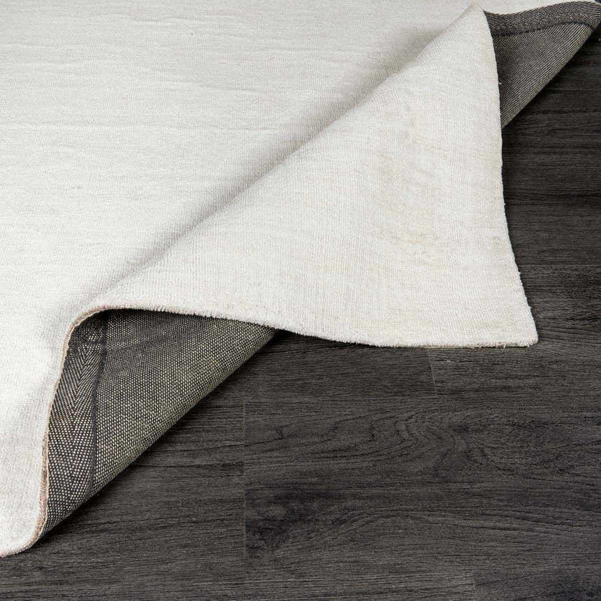 Close-up of the white, hand-loomed Brownsdale Area Rug showing its texture and backing on dark wood floors. Fold