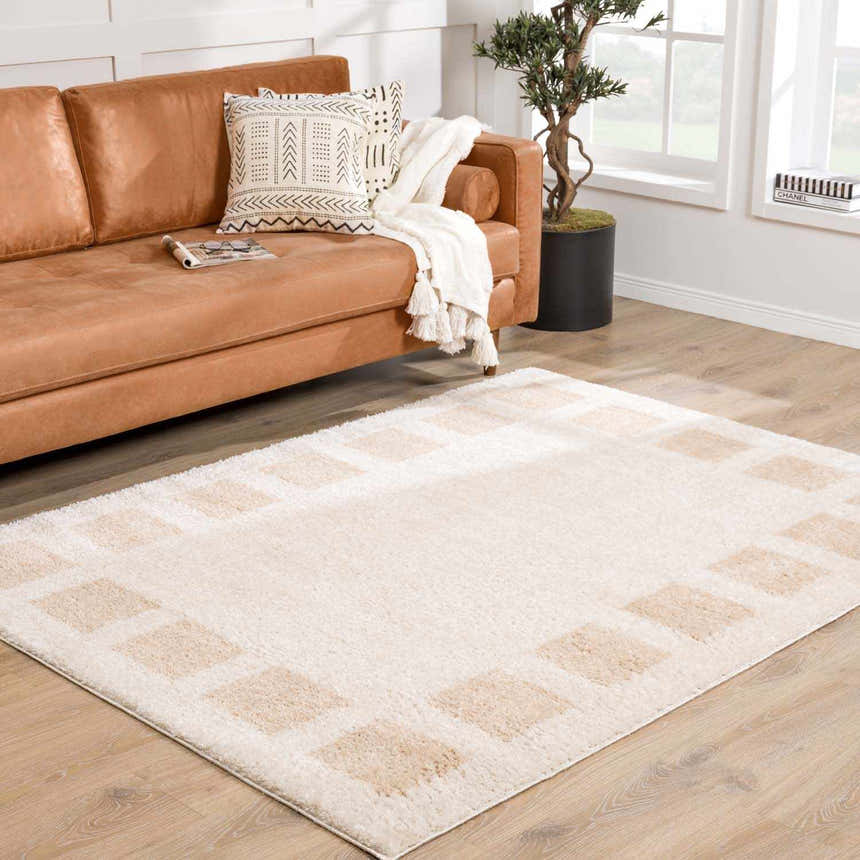 Living room scene with a Beynon Beige Washable Area Rug, tan leather sofa, and light wood floors. Roomscene-Rectangle