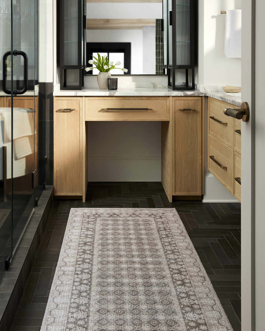 Bathroom scene with a Rainier Light Brown Washable Rug, light wood vanity, black herringbone tile floor, and glass shower. Roomscene-Runner