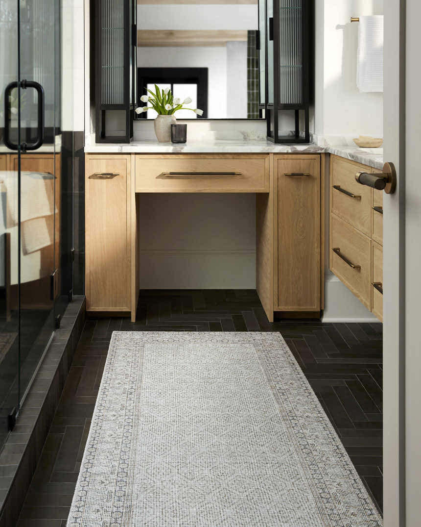 Bathroom scene with a Rainier Cottage Border Beige Washable Rug, wood vanity, and dark herringbone tile floor. Roomscene-Runner