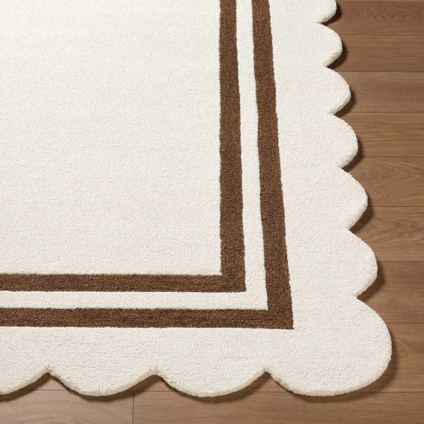 Close-up of the hand-tufted Chogan Ivory Area Rug. Features a brown border and a scalloped edge on a wood floor. Front