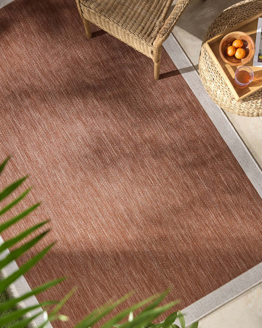 Close-up of a Chetan Brown outdoor rug with a woven texture and cream border, styled on a patio with wicker furniture. Roomscene-Rectangle