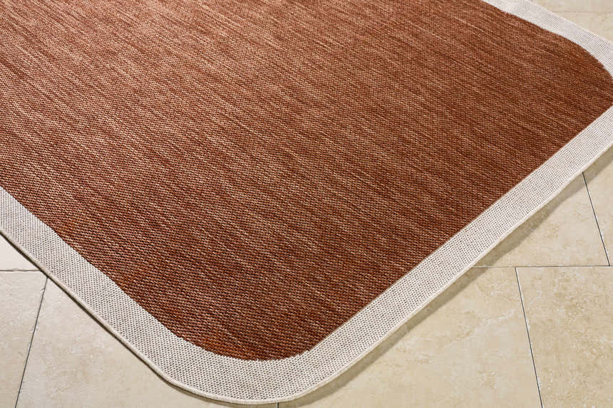 Close-up of a Chetan Brown Outdoor Rug showing its woven texture and cream border on a tile floor. Corner