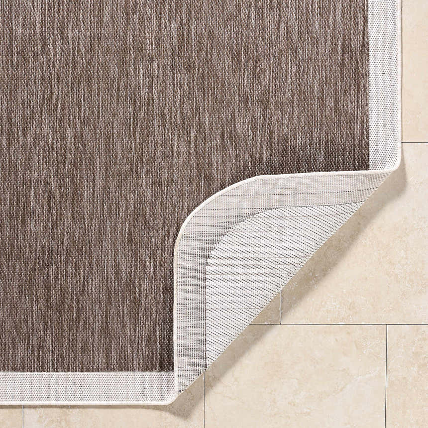 Close-up of the Chetan Dark Brown Area Rug showing texture, cream border, and non-slip backing on beige tile. Fold