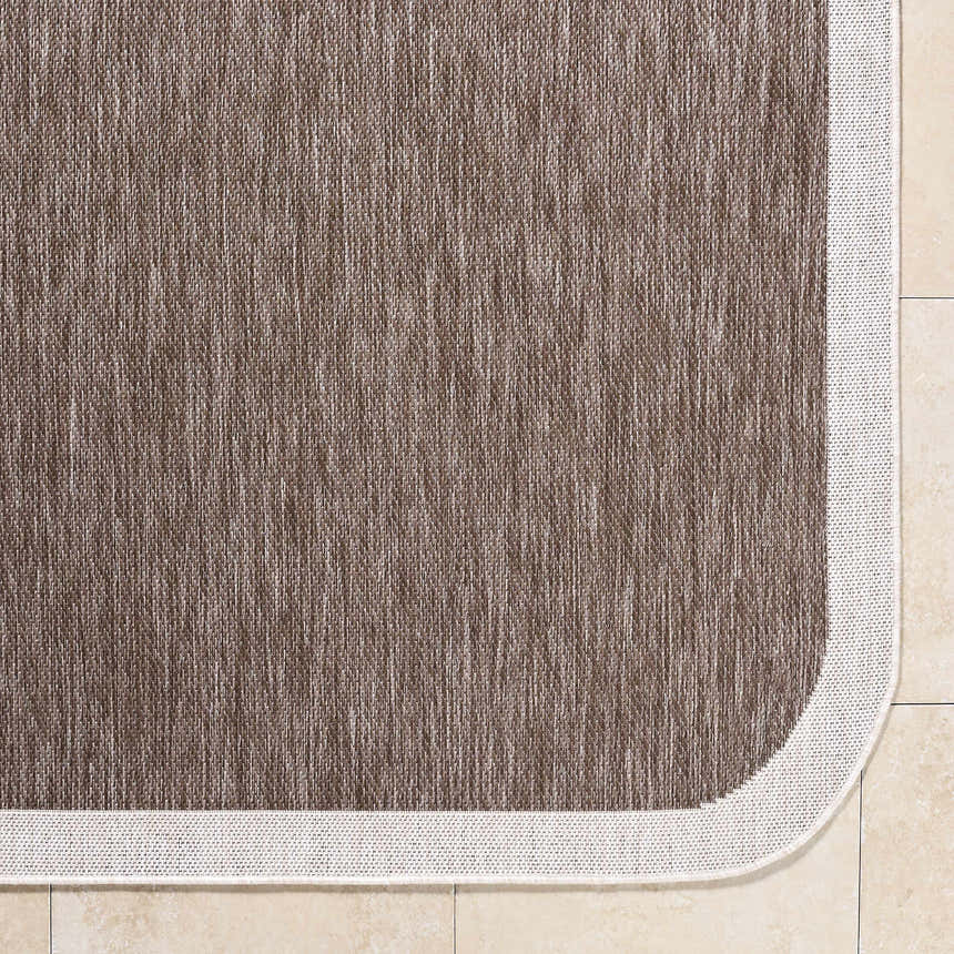 Close-up of the Chetan Dark Brown Area Rug showing its woven texture and cream border on beige tile. Corner