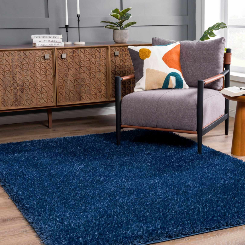 Living room scene with a Faina Solid Dark Blue Shag rug, gray chair with colorful pillow, and a wood cabinet. Roomscene-Rectangle