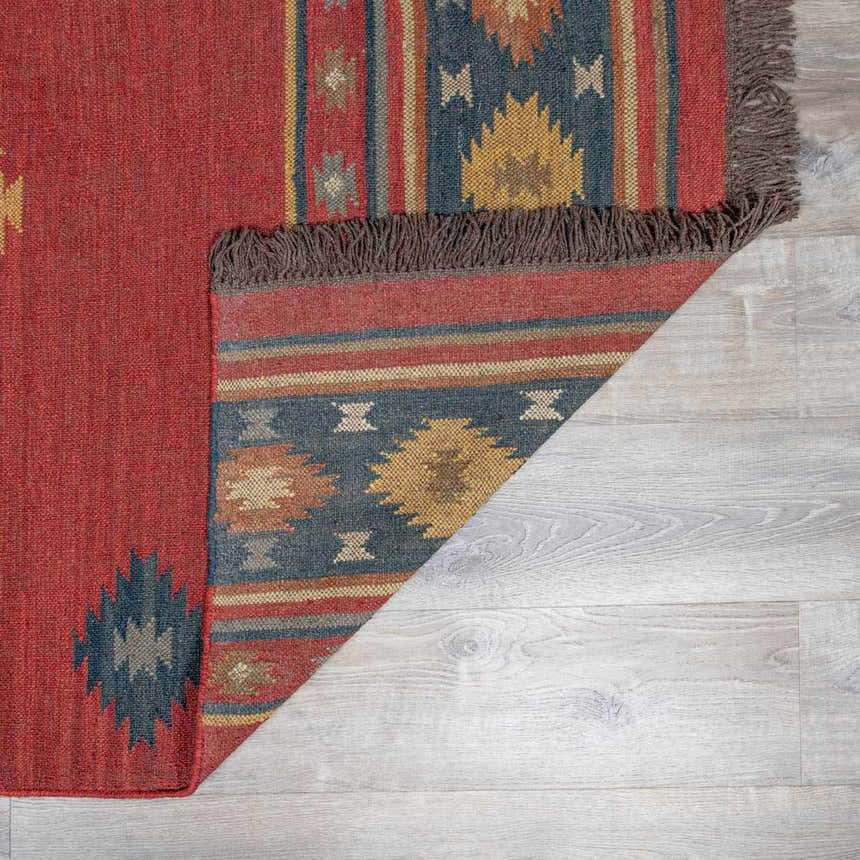 Close-up of the Hedon Wool Area Rug. Brick red rug with southwestern-style geometric patterns in blue, tan, and brown. Fringe detail. Fold