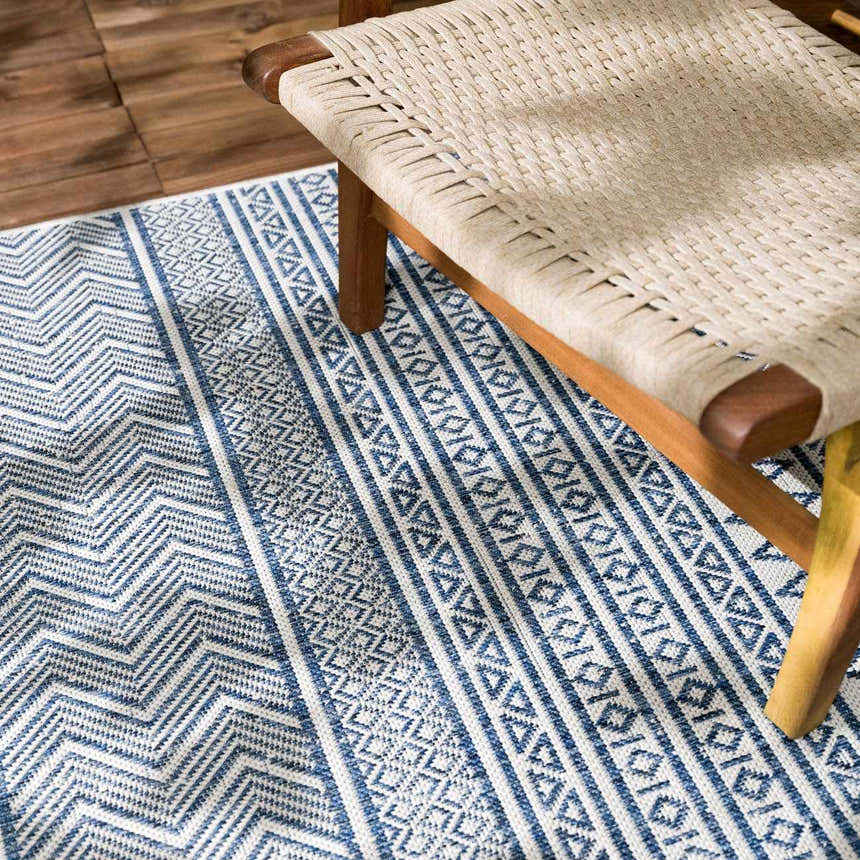 Close-up of the Madras Light Blue Green All Over Performance Area Rug with blue and white geometric patterns, next to a woven chair. Styleshot