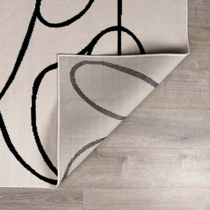 Close-up of the Eilir Modern Boho Rug showing bone color, black abstract design, and rug backing on light wood floor. Fold