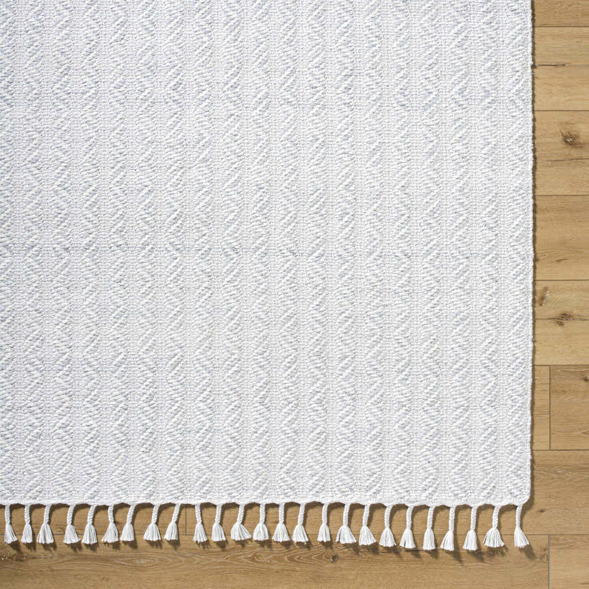 Close-up of the Anelia Powder Blue Area Rug. Hand-woven rug with ivory and powder blue diamond pattern and tassel fringe. Corner