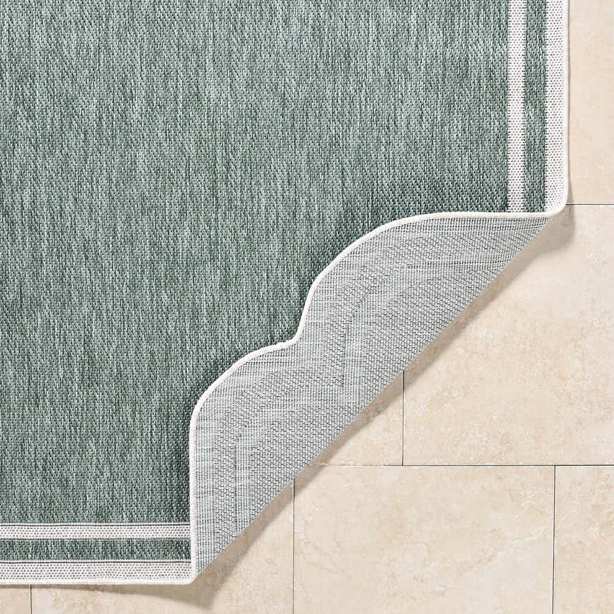 Close-up of the Arnola Green Outdoor Rug showing its texture, cream border, and non-slip backing on beige tile. Fold