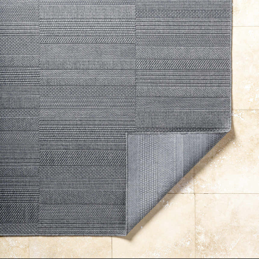 Close-up of the Thana Gray Area Rug showing its charcoal color, woven texture, and geometric pattern on a tiled floor. Fold