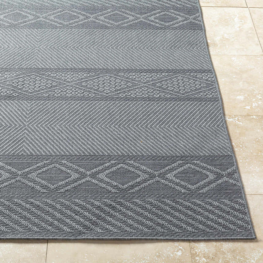 Close-up of the gray Aletha Area Rug showing its woven geometric pattern and texture on a beige tile floor. Front