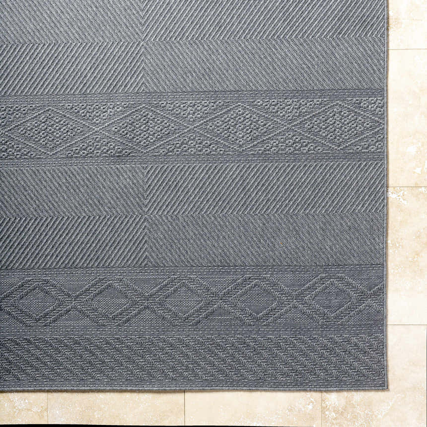 Close-up of the gray Aletha Area Rug showing geometric patterns and woven texture on a beige tile floor. Corner