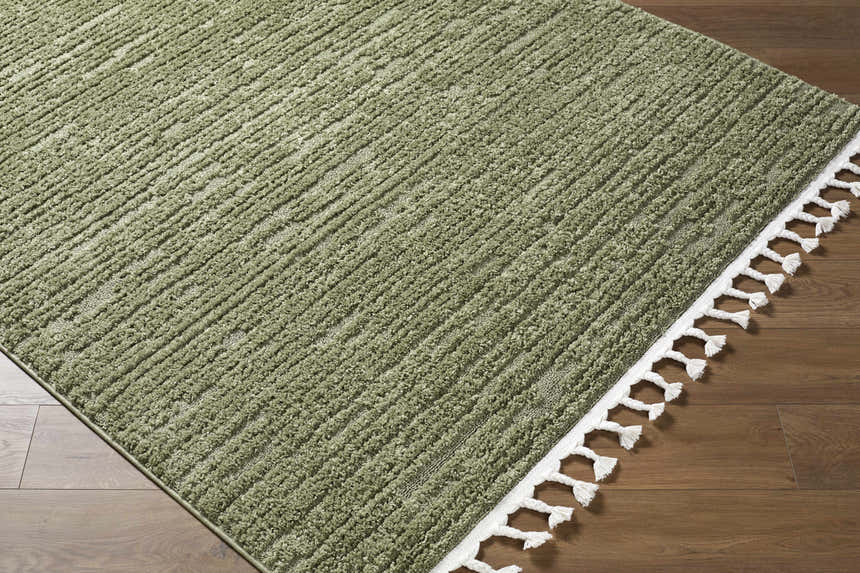 Close-up of the olive Annily Area Rug, a machine-woven rug with a subtle stripe texture and white tassels on a wood floor. Corner