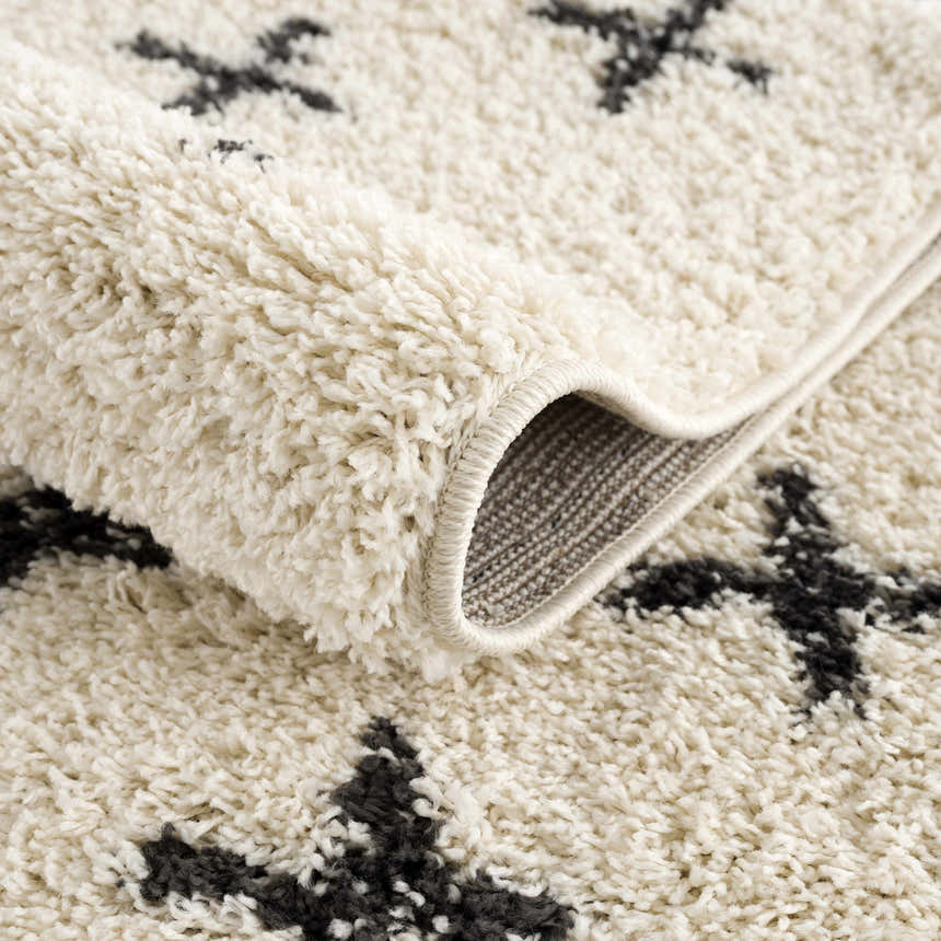 Close-up of the Alstead Shag Area Rug, showing its beige shag texture and charcoal Moroccan-inspired cross pattern. Fold