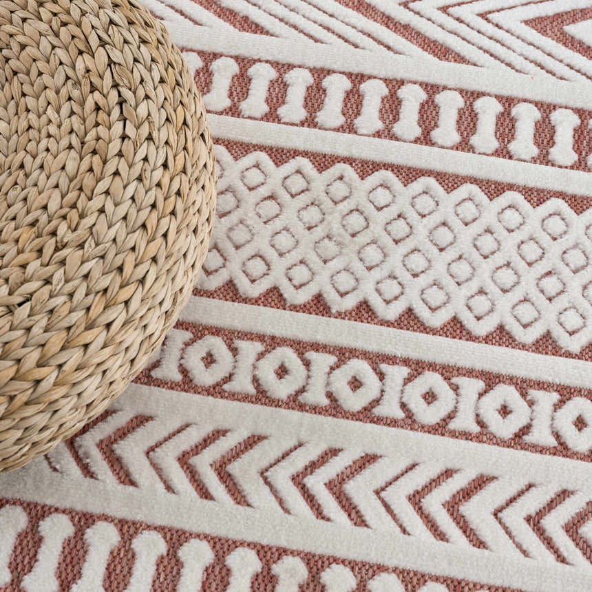 Close-up of Jove Orange Outdoor Rug showing bone white geometric pattern on terracotta, with a woven ottoman. Styleshot