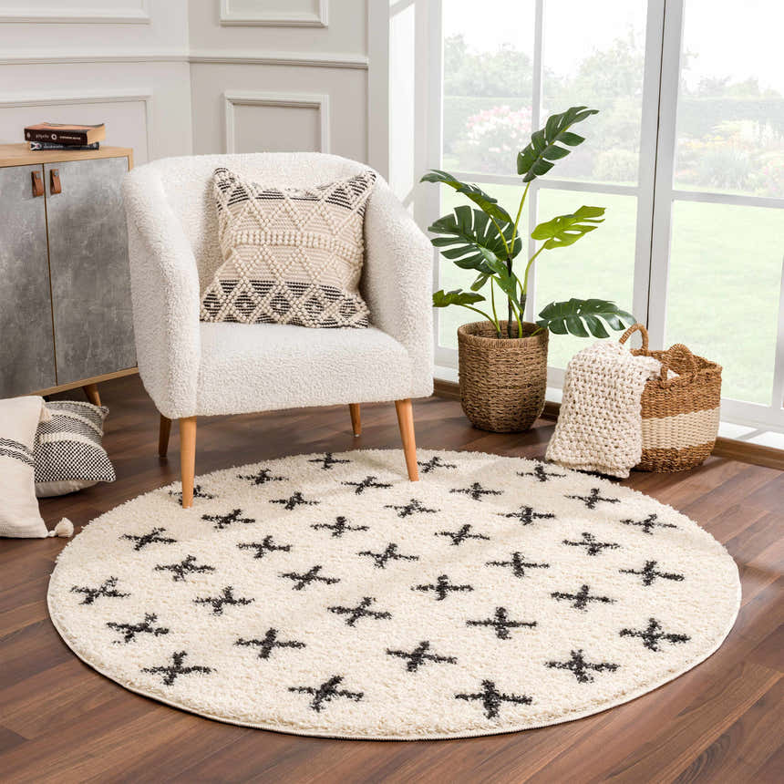 Cozy living room scene with a round, beige Alstead Shag Area Rug featuring a black plus sign pattern under a bouclé chair. Roomscene-Round
