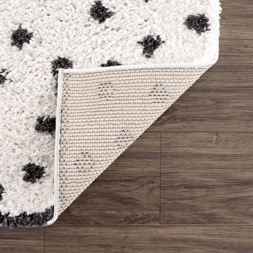 Close-up of the Chaia Dotted Black & White Plush Rug showing texture, black dots, and the rug's backing on a wood floor. Fold