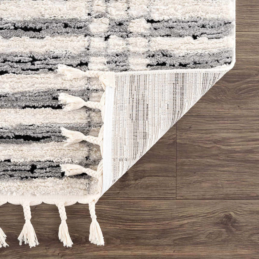 Close-up of the Nava Area Rug showing beige, black, and bone geometric pattern, tassels, and machine-woven construction. Fold