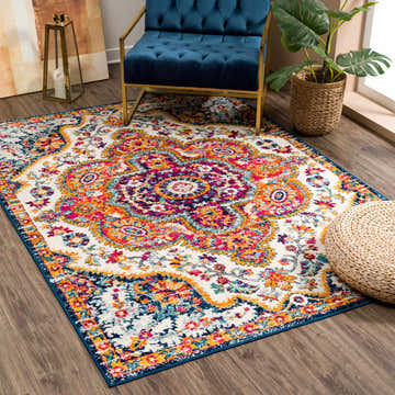 Living room scene with a colorful New Simsbury Area Rug, blue velvet chair, and woven pouf on a wood floor. Roomscene-Rectangle