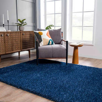 Living room scene with Faina Solid Dark Blue Shag rug, gray chair, patterned pillow, and wooden accent table. Roomscene-Rectangle