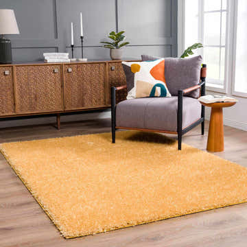 Living room scene with Faina Solid Gold Shag Washable Area Rug, gray chair, wood cabinet, and modern decor. Roomscene-Rectangle