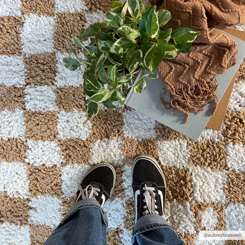 Close-up of the Kieu Mustard & Ivory Checkered Area Rug with a plant, book, scarf, and person wearing jeans and sneakers. Roomscene-Rectangle