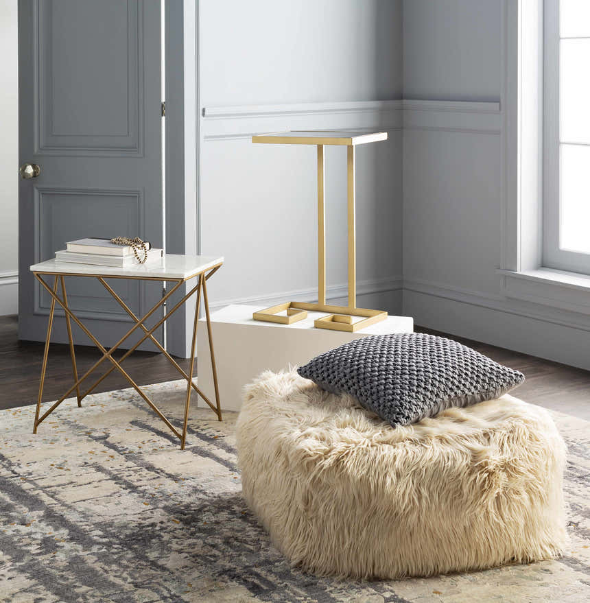 Contemporary gray room scene with a Moncure Area Rug, gold side tables, and a fluffy beige ottoman. Styleshot