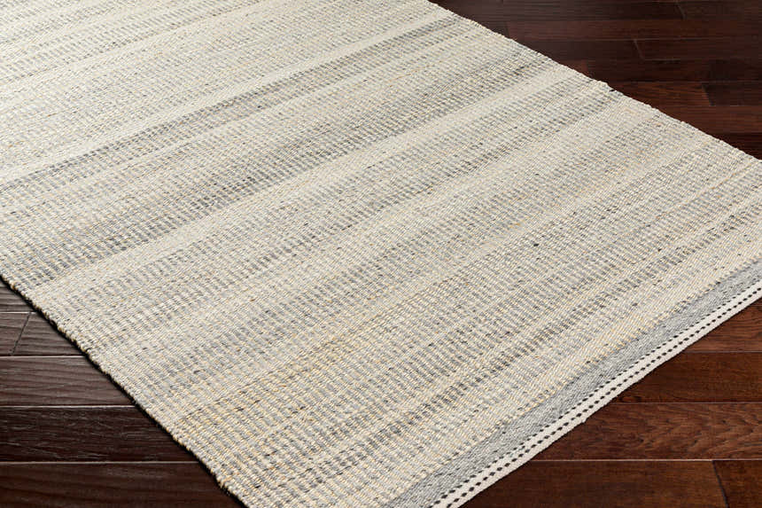 Close-up of the Abucay Jute Rug. Hand-woven rug in charcoal, cream, light beige, and light gray on a dark wood floor. Corner