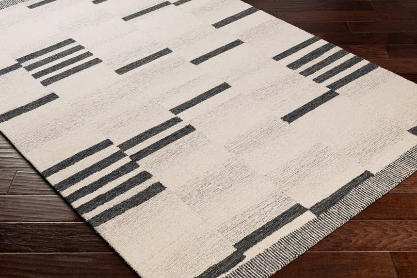 Close up of Aibonito Hand Tufted Wool Area Rug. Beige rug with black geometric stripes on a dark wood floor. Corner