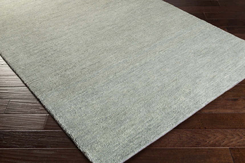 Close-up of the sage Ammanford Area Carpet showing its hand woven texture on dark wood floors. Corner