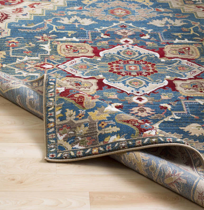 Close-up of the Argyle Area Rug showing the traditional pattern in brick red, dark blue, and tan on a wood floor. Fold