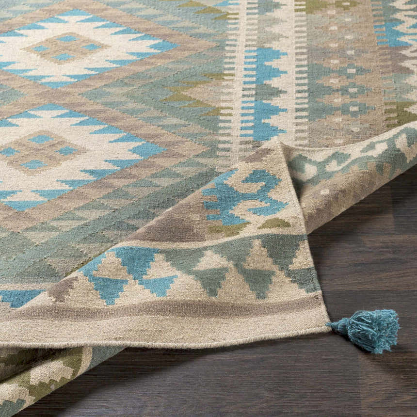 Close-up of the hand woven Auchterarder Carpet. Farmhouse style rug in aqua, blue, sage, and brown with geometric pattern and tassel. Fold