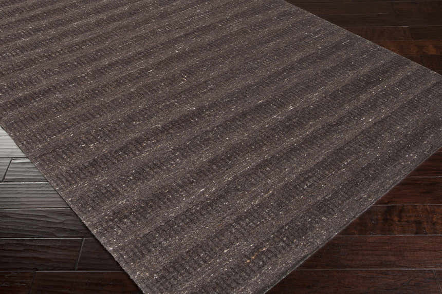 Close up of the hand-loomed Banbury Area Rug, a black wool rug with subtle stripes on dark wood flooring. Corner