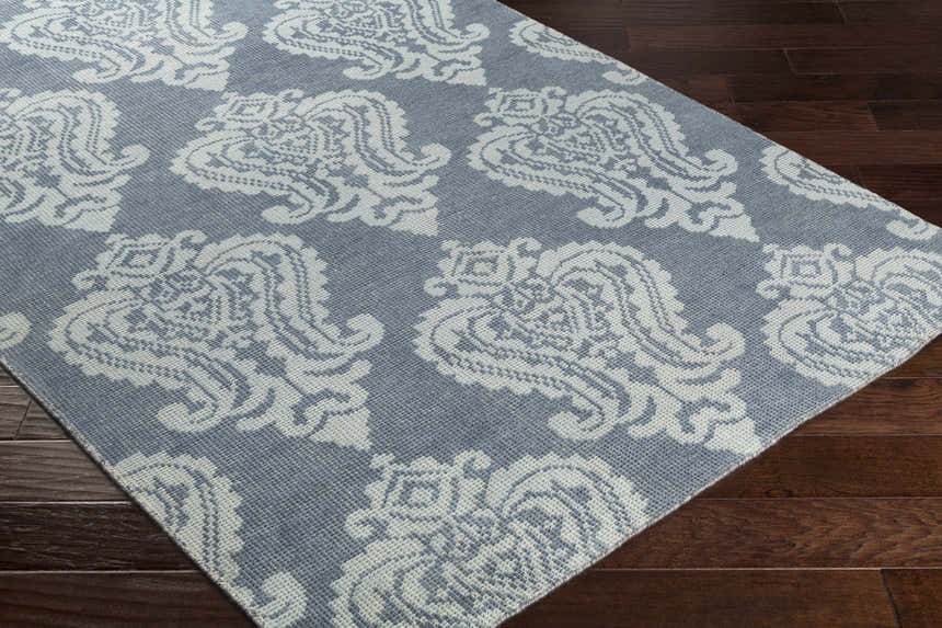 Close-up of the Bargersville Area Carpet. Blue wool rug with a pale blue traditional damask pattern on dark wood floors. Corner