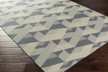 Close-up of the Bayard Area Carpet. Hand-woven wool rug with a geometric pattern in denim, ivory, light sage, and medium gray. Corner
