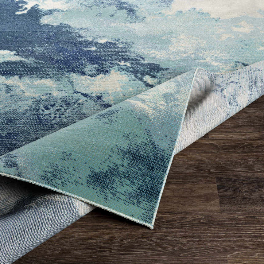 Close-up of Bright Sky Indoor/Outdoor Rug showing ivory, light slate, navy, and pale blue abstract pattern on dark wood floor. Fold
