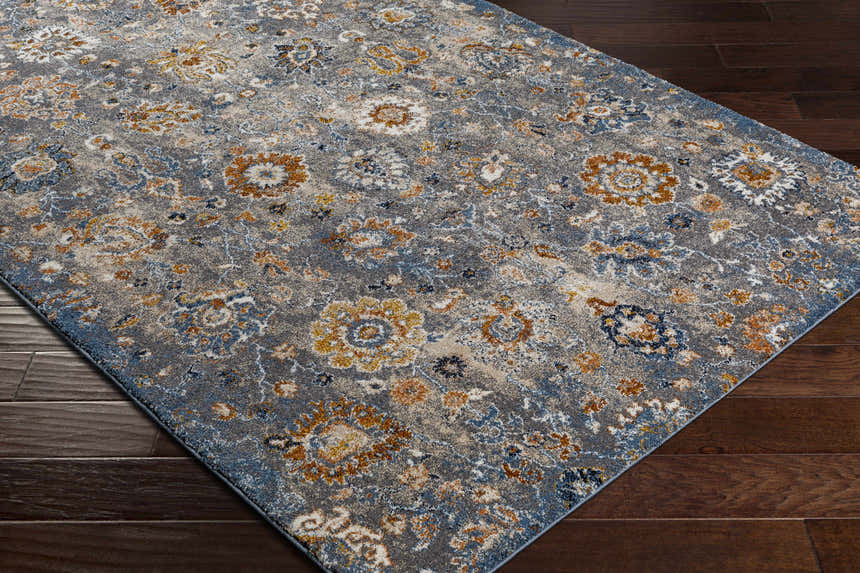 Close-up of the Cogan Gray Thick Rug. Features a floral pattern in shades of gray, blue, cream, and brown on dark wood floors. Corner