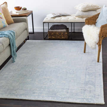 Living room scene with a Calderbank Sky Blue wool-blend area rug, gray sofa, white bench, and woven chair on dark wood floors. Roomscene-Rectangle