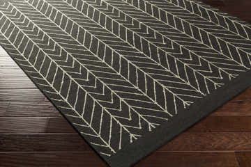 Close-up of the Clymer Wool Rug. Black rug with off-white geometric herringbone pattern on dark wood floors. Corner