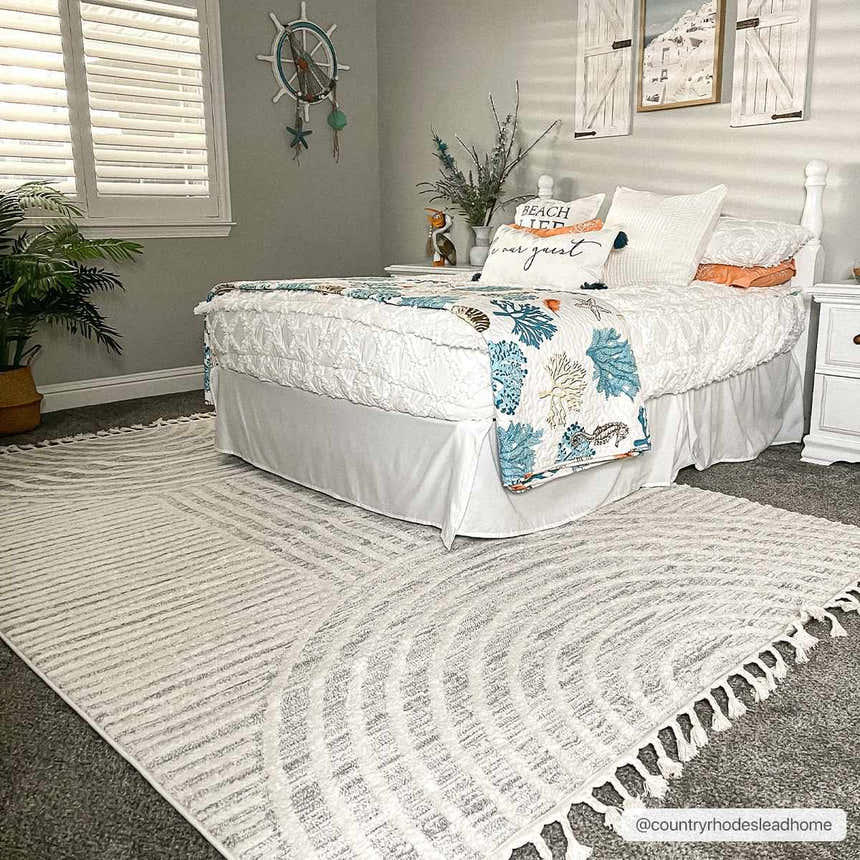 Coastal bedroom scene with a Dauin 3D Rainbow Area Rug in cloud gray, cream, and gray tones with tassels. Roomscene-Rectangle