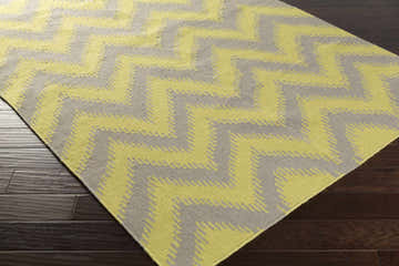 Close-up of the hand woven Cramlington Area Rug. Gray and light olive chevron pattern. Wool rug on dark wood floor. Corner