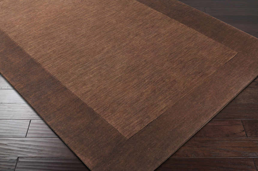 Close-up of a Bordered Solid Brown Wool Rug showing texture and color variations on dark wood floor. Corner