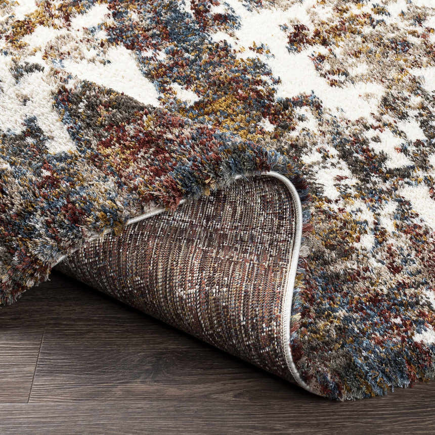 Close-up of the Darenth Area Rug. Shag texture with burgundy, cream, blue, and mustard hues on a dark wood floor. Fold